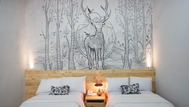 A double bed with white bedding and patterned pillows, a wooden nightstand with a lamp, and a wall mural of a deer in a forest. - Omah Njonja Bed & Brasserie (Depok)