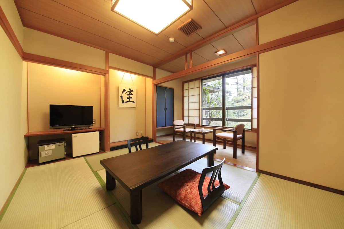 Um quarto japonês tradicional com tatames, uma mesa baixa, cadeiras, uma televisão e uma porta deslizante que dá para uma janela com vista para árvores.