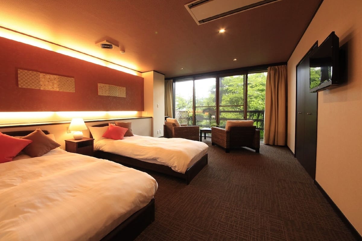 Quarto de hotel com duas camas, televisão, varanda com vista para o verde e chão coberto de carpete.