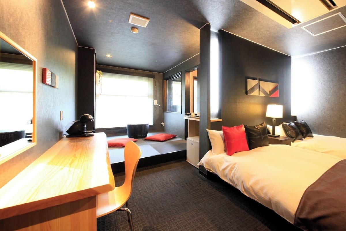 Quarto de hotel moderno com cama, escrivaninha e área de estar junto à janela.