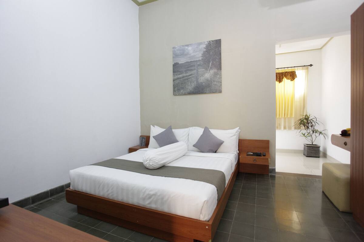 Un dormitorio ordenado con cama, mesitas de noche, una planta en maceta y un cuadro en la pared.