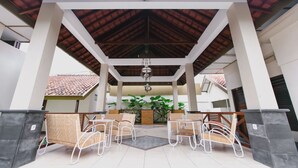 Un espacio de patio al aire libre con sillas y mesas de metal blanco, techo de madera y vista a un edificio con tejado de tejas.