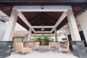 Un espacio de patio al aire libre con sillas y mesas de metal blanco, techo de madera y vista a un edificio con tejado de tejas.