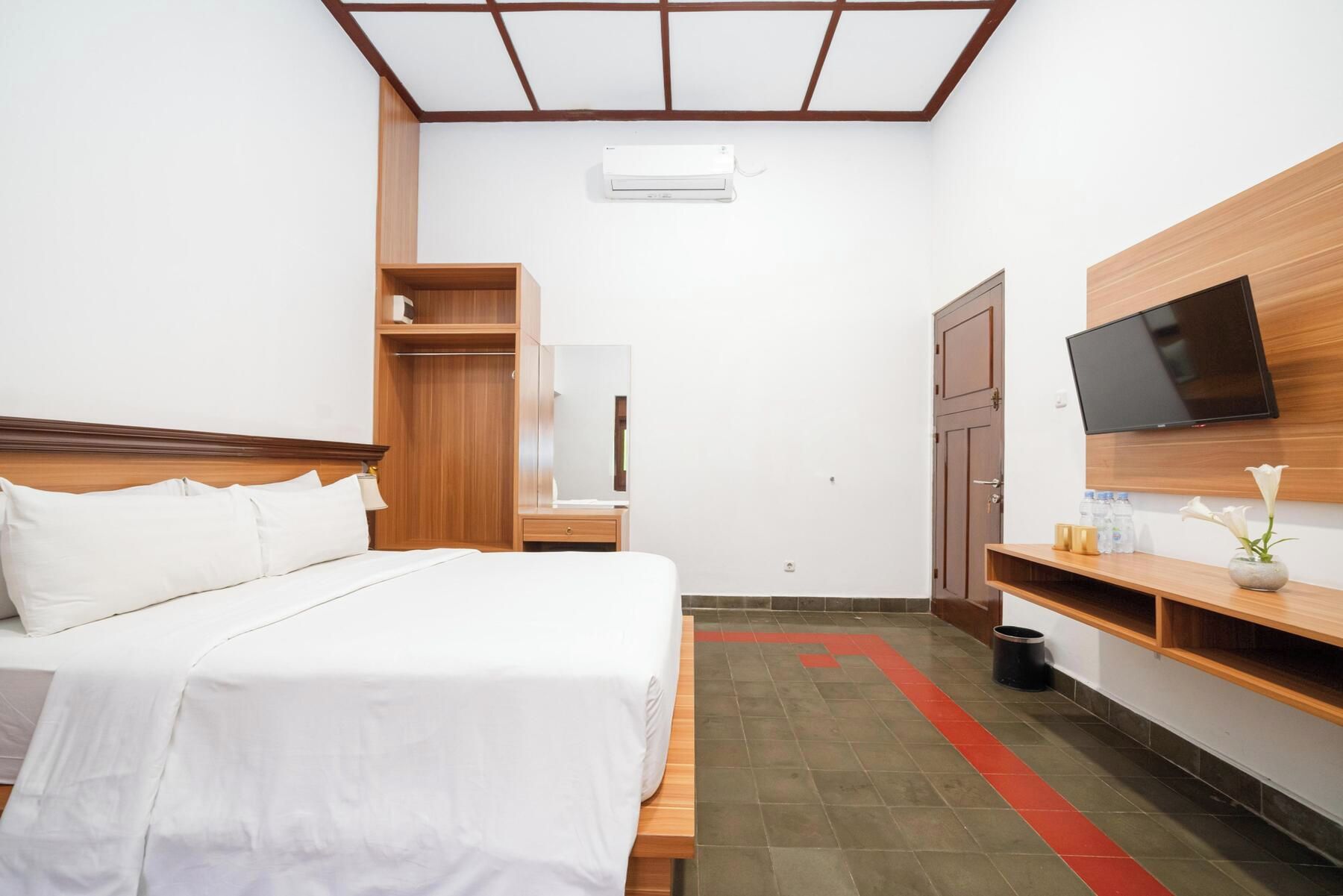 A hotel room with a bed, a wardrobe, a television mounted on the wall, and a small table with a vase of flowers.