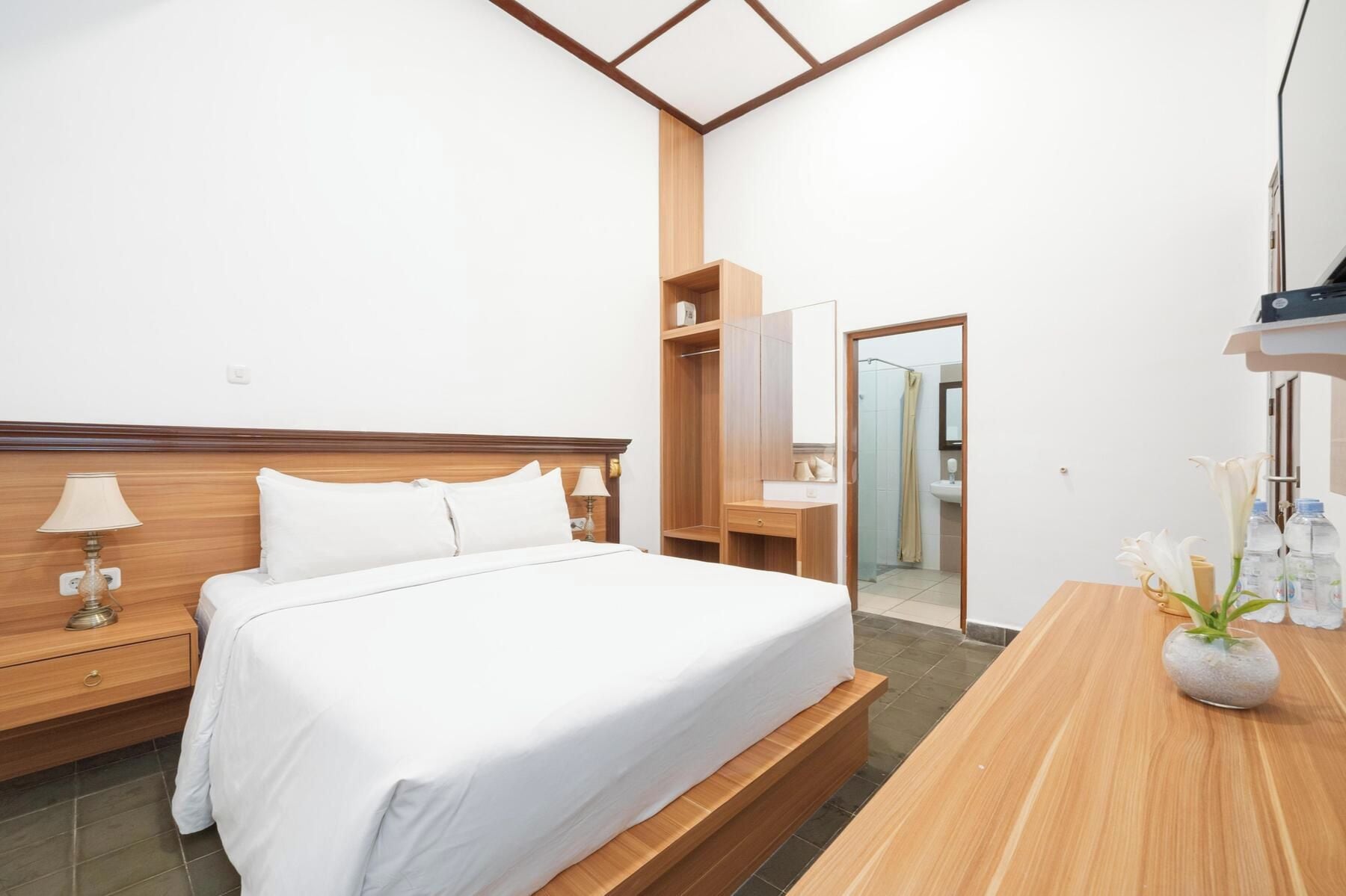 A hotel room with a bed, bedside table, lamp, wardrobe, and a bathroom visible through a glass door.