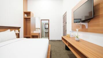 Quarto de hotel com cama, escrivaninha de madeira, televisão presa à parede e banheiro visível por uma porta aberta.