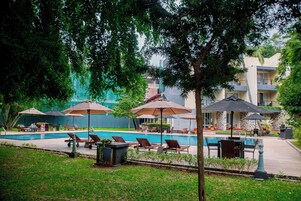 Zona de piscina con sillones de descanso y sombrillas, rodeada de vegetación y un edificio al fondo.