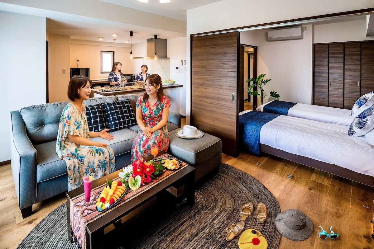 Een moderne hotelkamer met een bank, een eettafel met fruit en snacks, een bed met blauw beddengoed, en een keuken in de achtergrond.