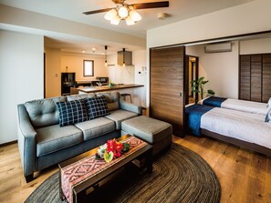 A modern living room with a sofa, a coffee table with flowers, a bed, and a kitchen in the background.