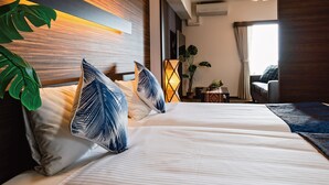 Ein modernes Hotelzimmer mit einem großen Bett, einem Holz-Kopfteil, einem Nachttisch, einer Topfpflanze und Blick auf das angrenzende Zimmer.