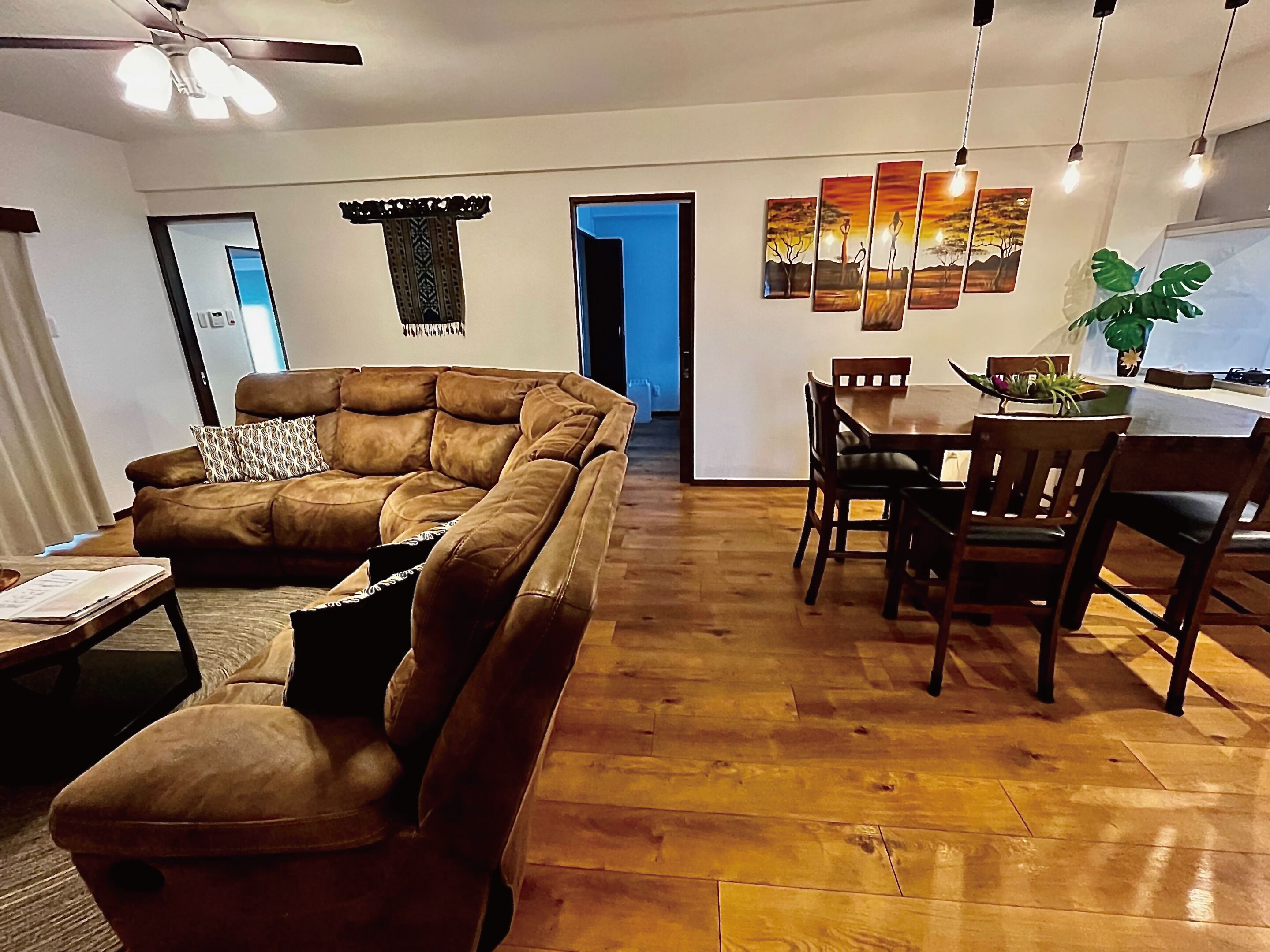 Una sala de estar con un sofá de cuero marrón, una mesa de comedor de madera y sillas. Hay un ventilador de techo, una obra de arte en la pared y una planta sobre la mesa de comedor.