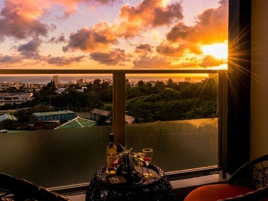 A room with a view of a sunset over a cityscape, a table with a bottle and glasses, and a chair.