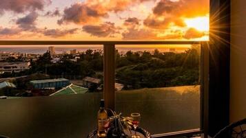 A room with a view of a sunset over a cityscape, a table with a bottle and glasses, and a chair.