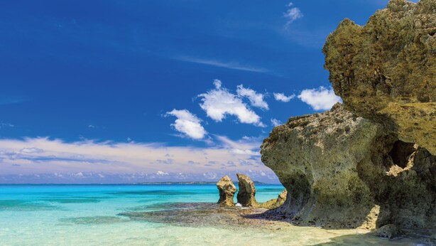 Ein Strand mit klarem türkisblauem Wasser, einem Sandufer und einer großen Felsformation mit einer hohlen Öffnung.