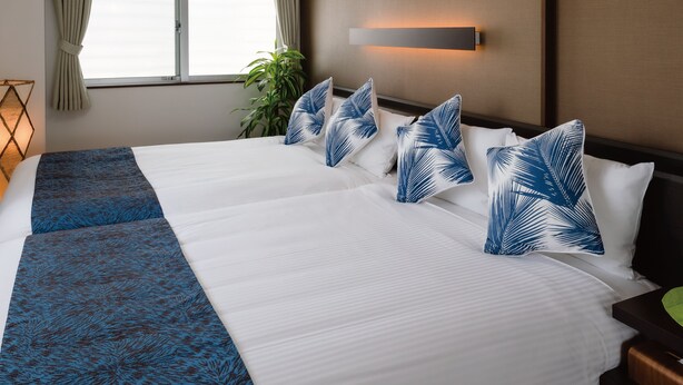 Una cama grande con ropa de cama blanca y azul, un plaid azul estampado y almohadones de hojas de palma.