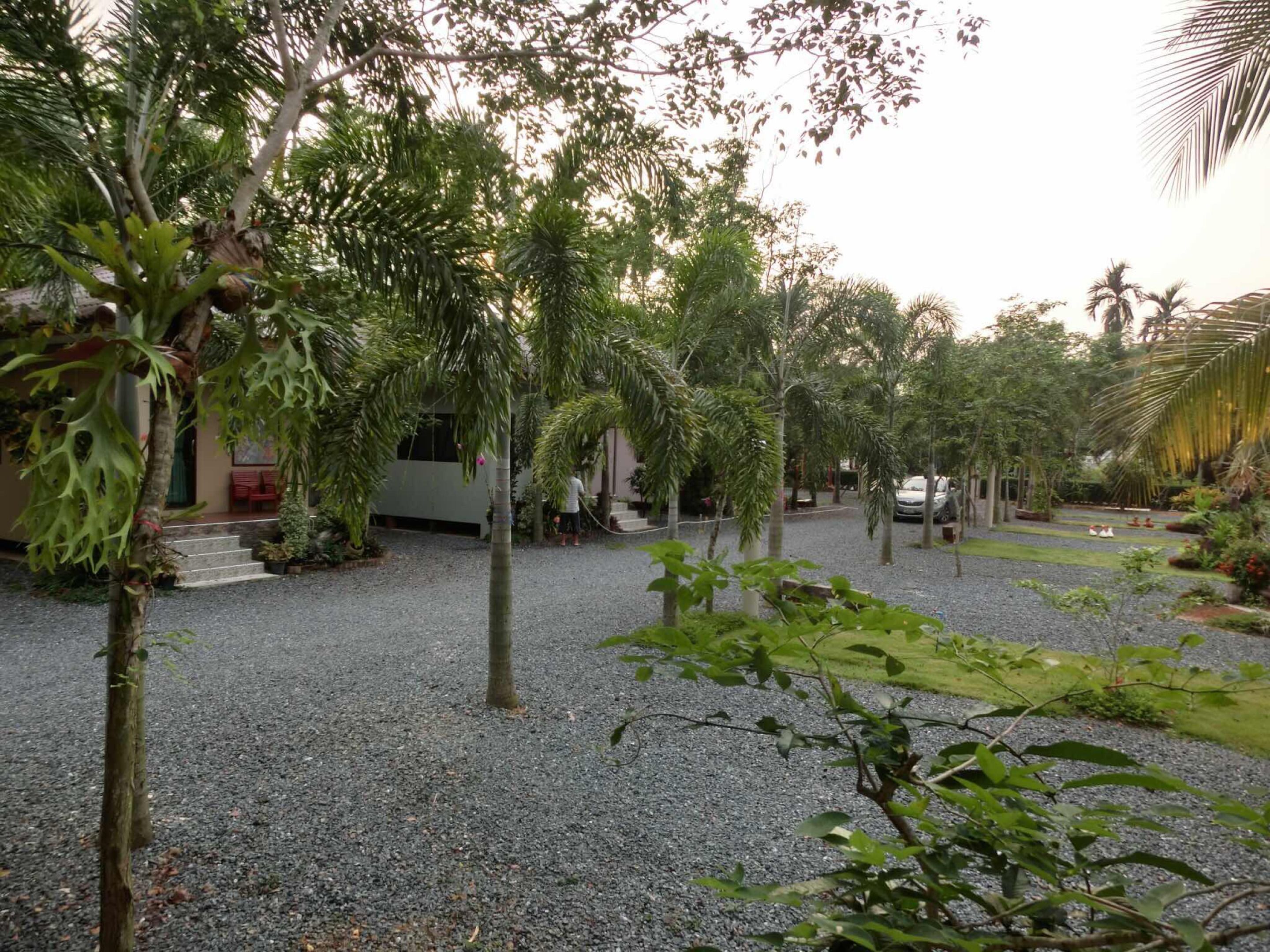 A courtyard with gravel ground, surrounded by various trees and plants, and a building with a red chair outside.