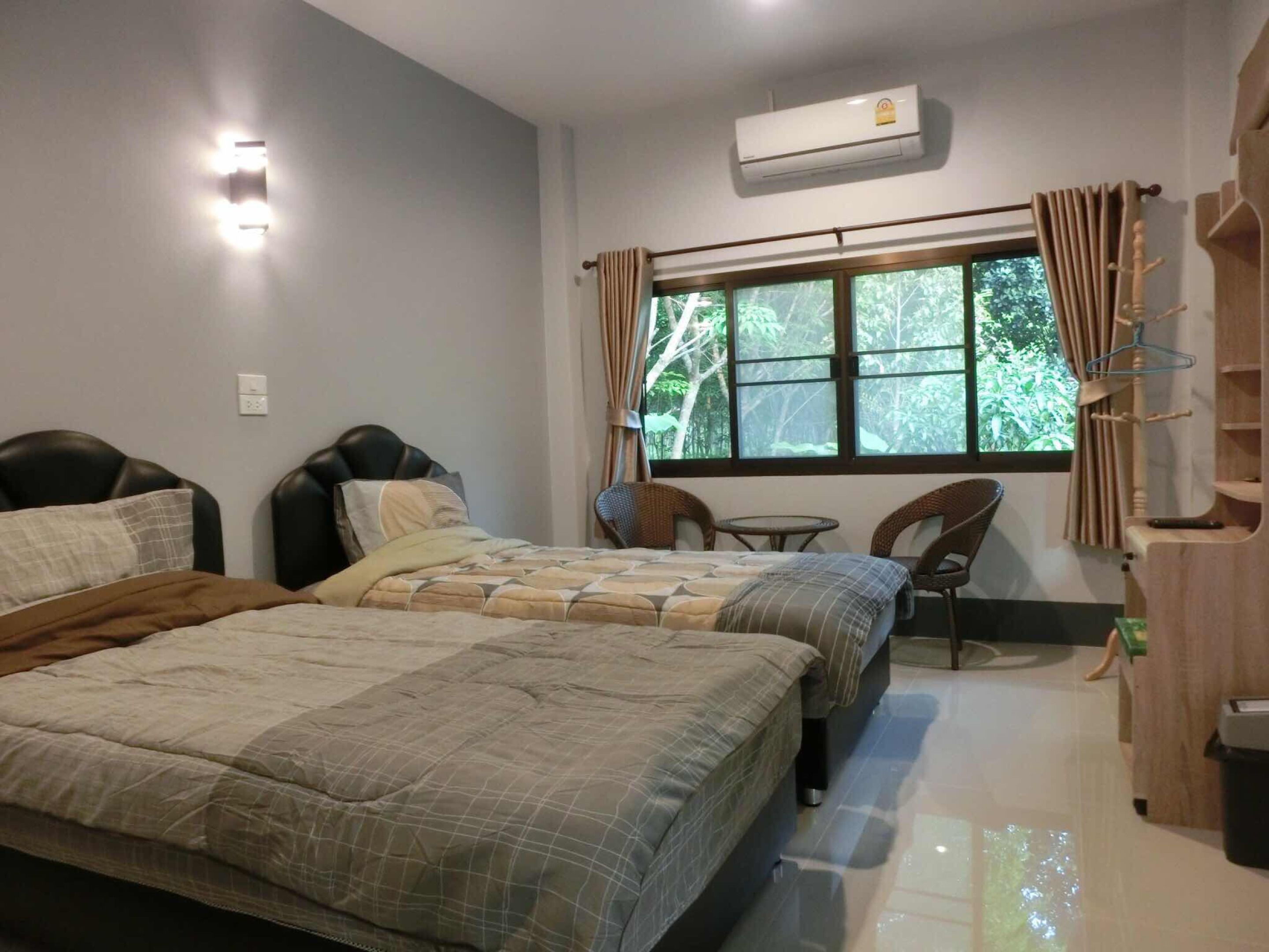 Une chambre d’hôtel avec deux lits, une petite table, des chaises, une fenêtre avec des rideaux, un climatiseur et une étagère en bois avec des cintres.