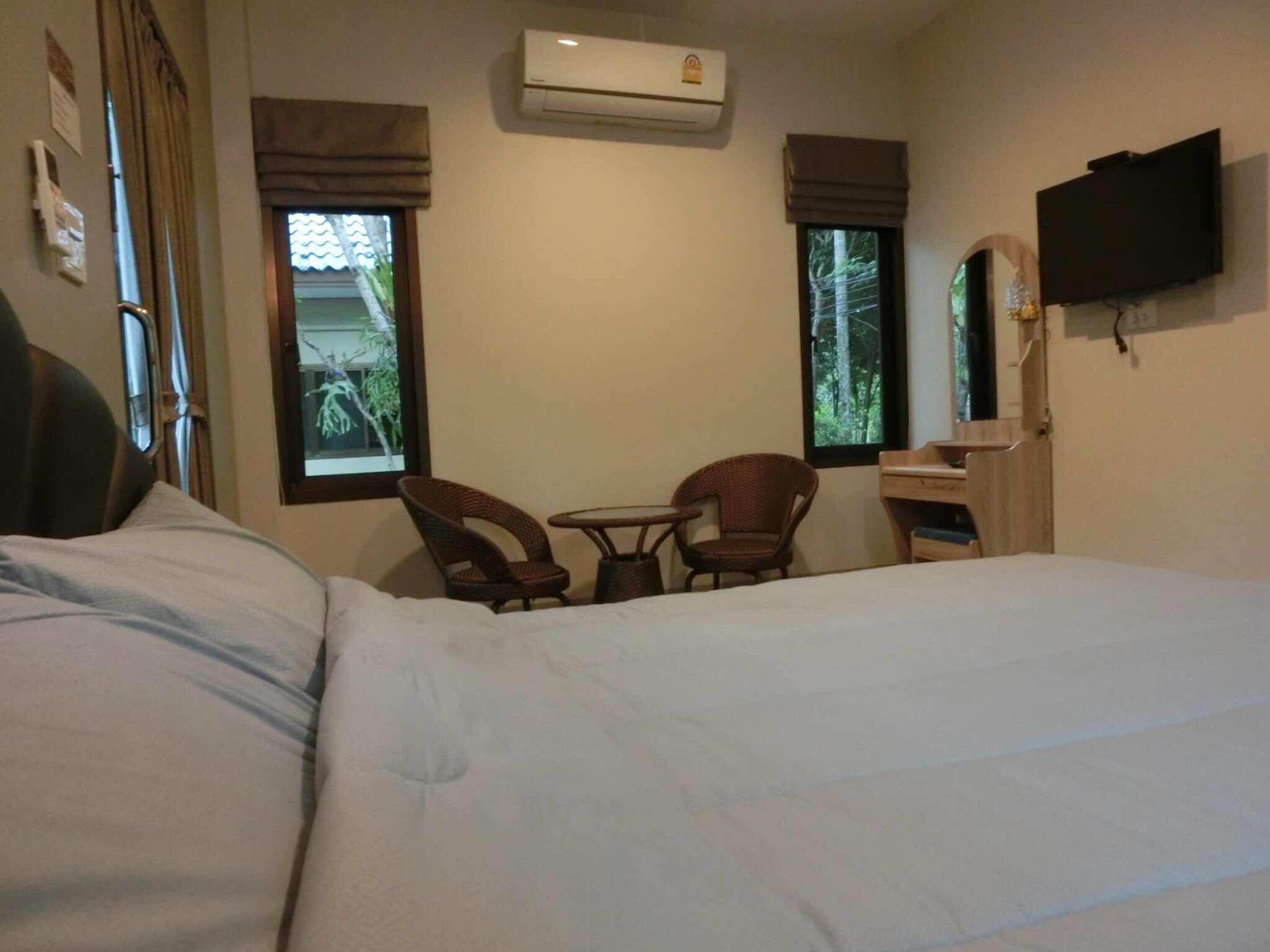 Hotelkamer met een bed, twee rieten stoelen, een tafeltje, een airconditioner, een televisie en ramen met gordijnen.