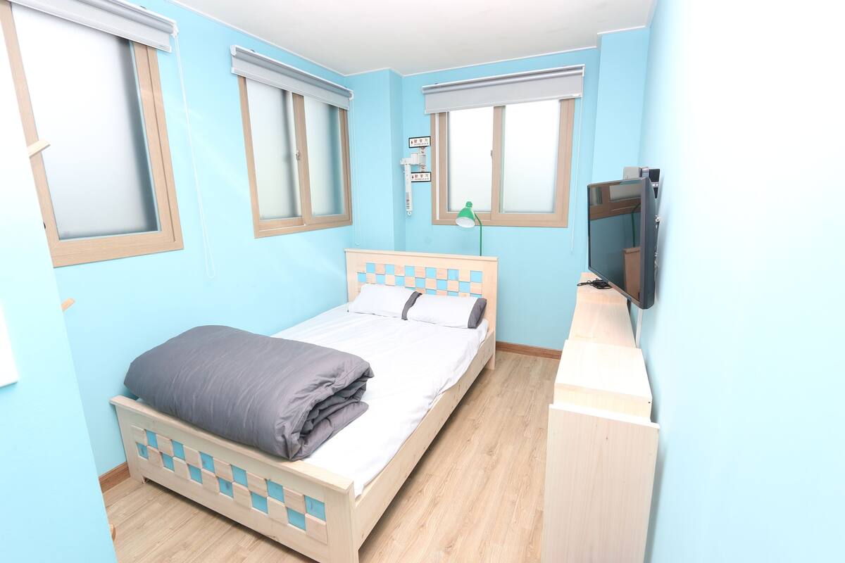 A bedroom with a bed, a TV, and two windows with blinds.