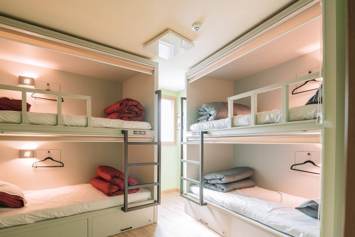 A dormitory room with bunk beds, each with a small shelf and a hanging rack.