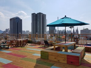 A rooftop terrace with wooden furniture, a blue umbrella, and a cityscape view.