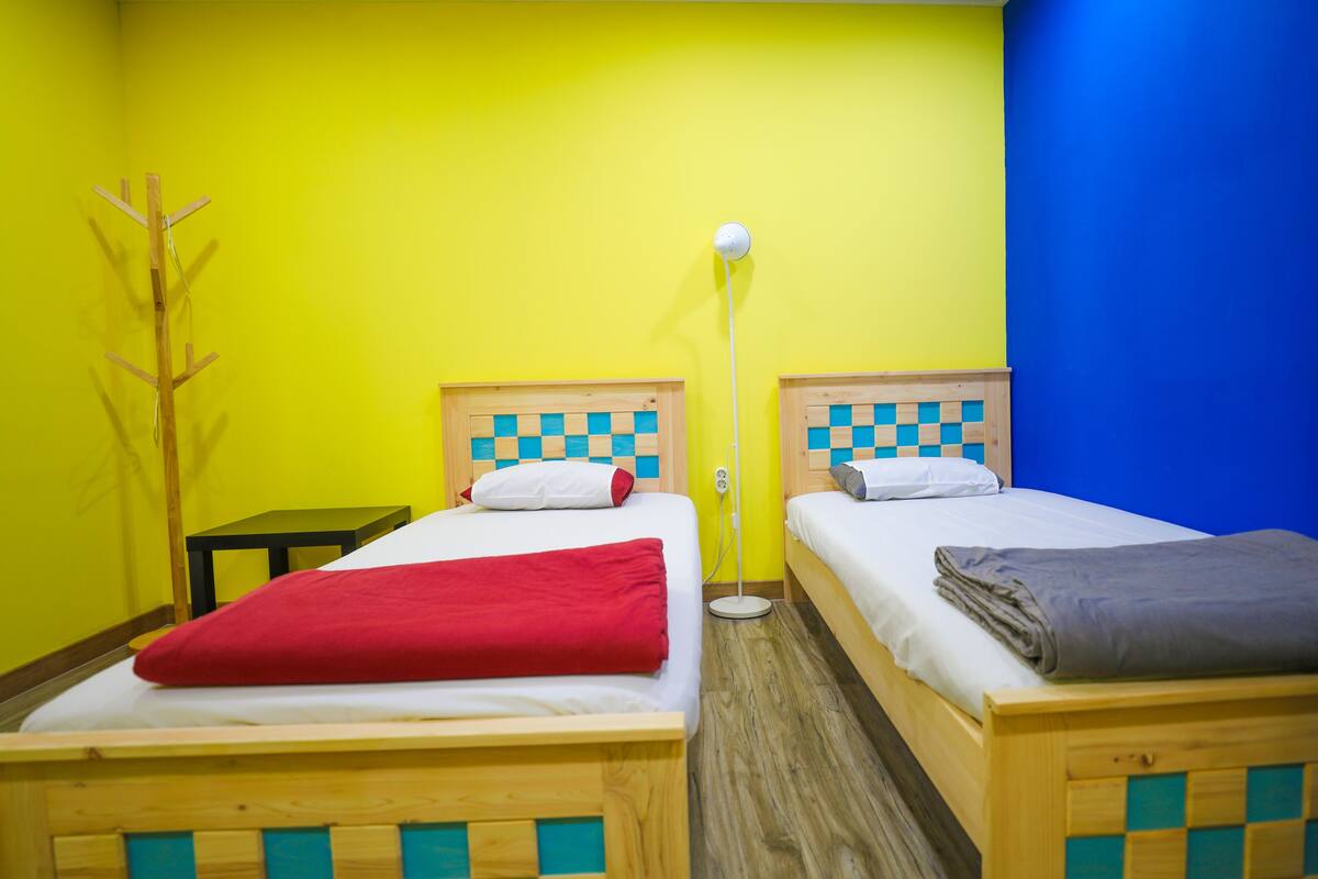 Two single beds with wooden frames and checkered headboards, a red blanket on one bed, a grey blanket on the other, and a floor lamp between them.