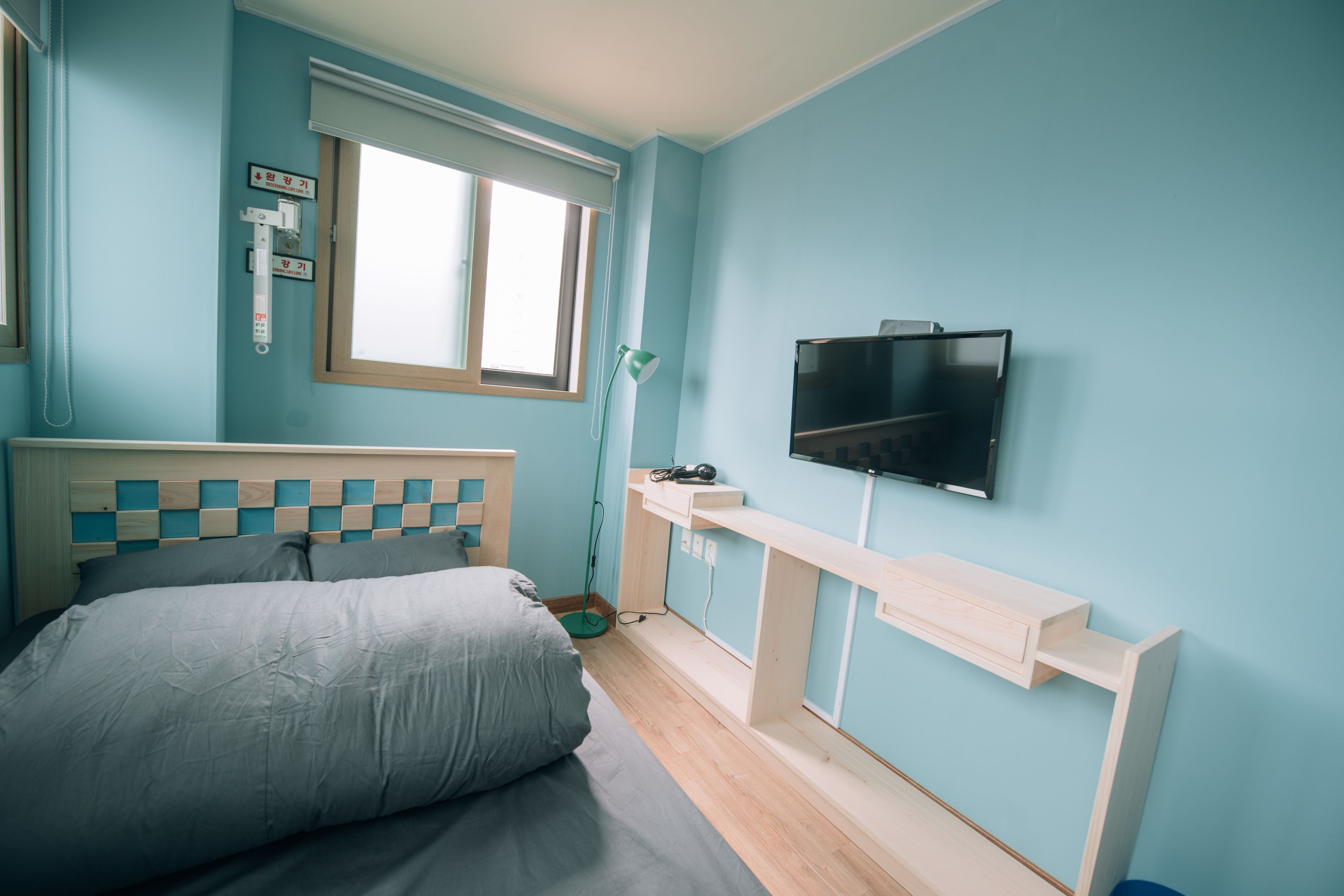 Un dormitorio con cama, televisor montado en la pared, ventana y mesita de noche.