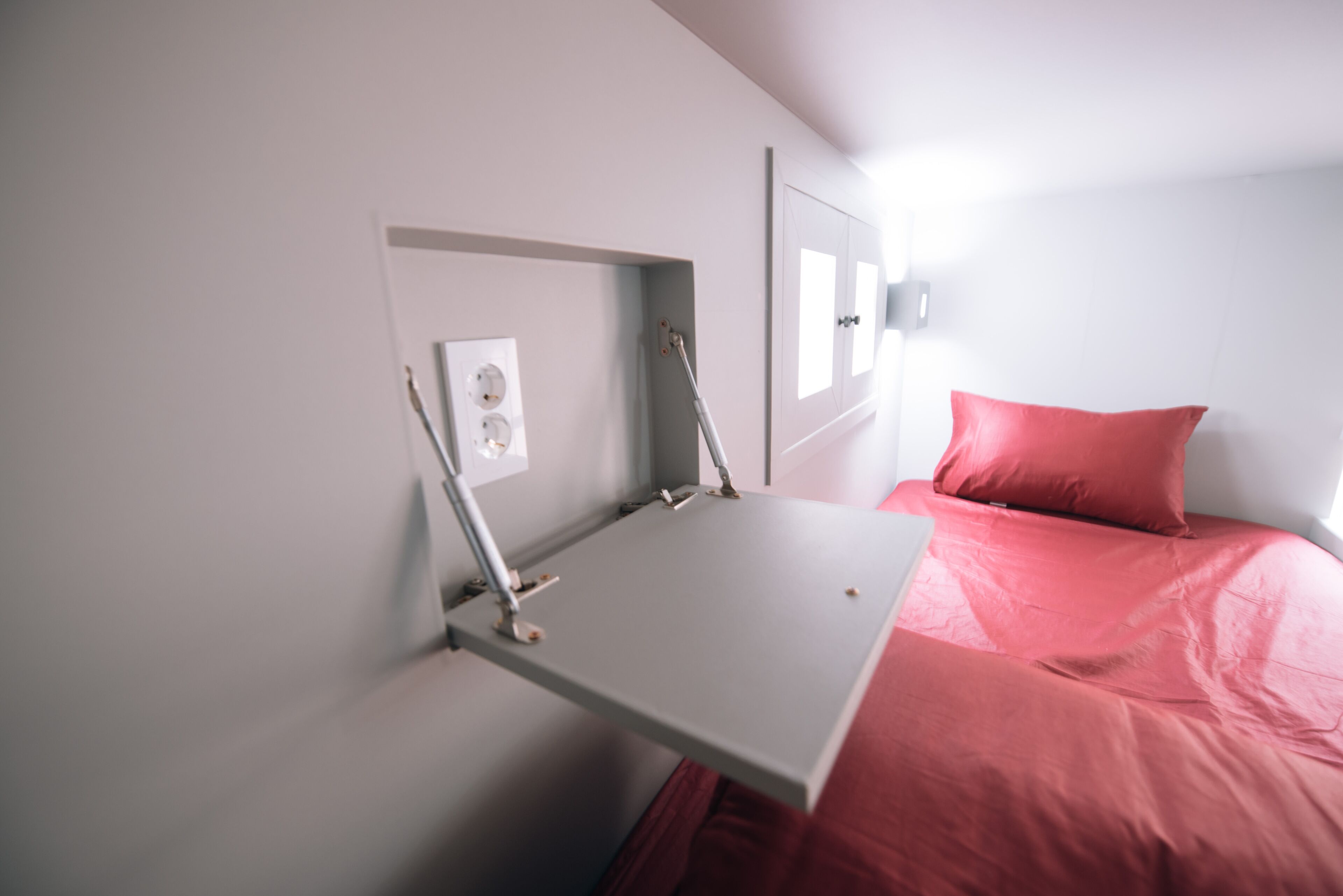 A compact room with a bed, a small desk, and a wall-mounted light fixture.