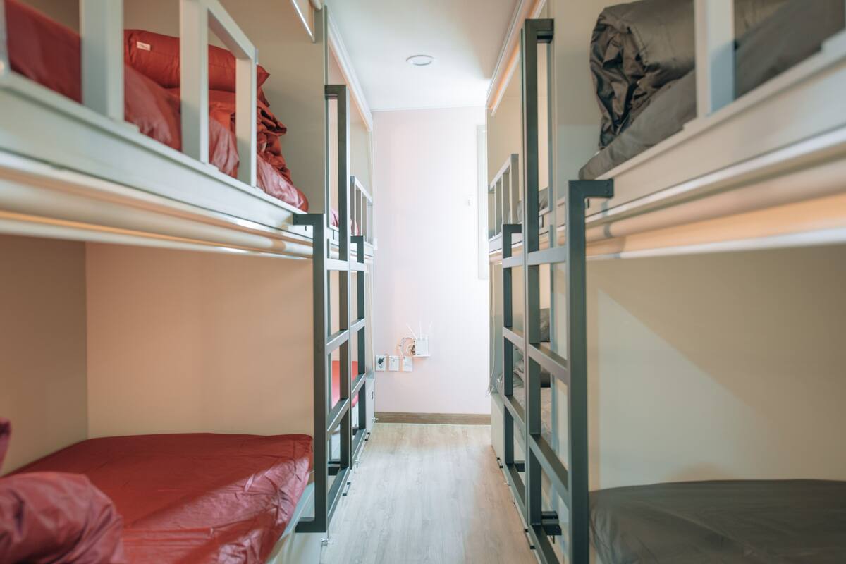 A narrow hallway with bunk beds on either side, a light-colored floor, and a small shelf with items on the wall.