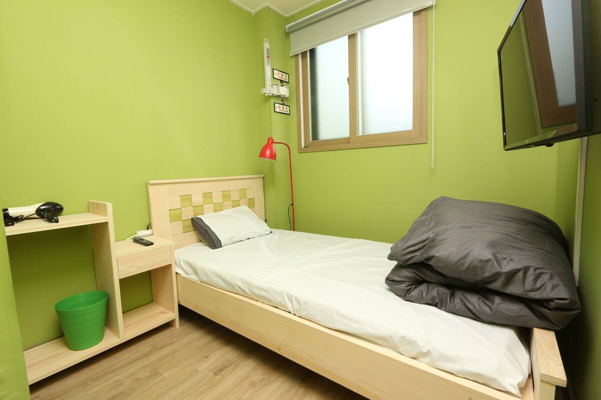 A small, single-bed room with a wooden bed, a green bucket, a red desk lamp, and a window with blinds.