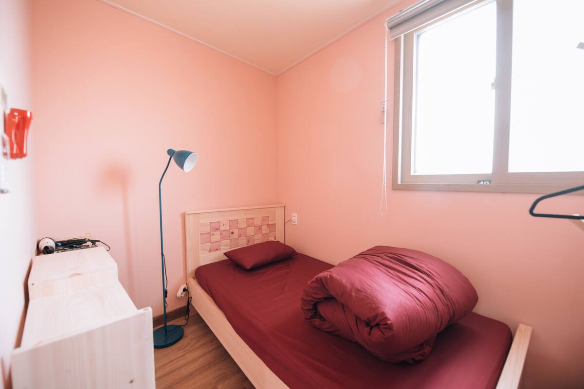 A small bedroom with a single bed, a bedside lamp, a window, and a wooden wardrobe.