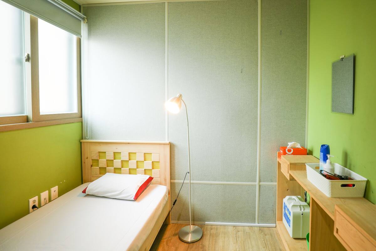 A room with a bed, a standing floor lamp, a small desk with a basket, and a wall-mounted shelf.