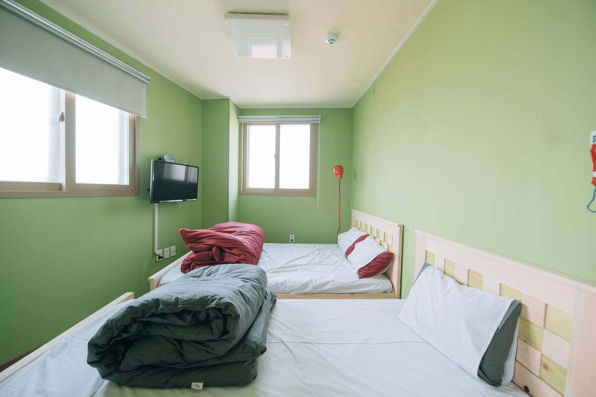 A room with two beds, a television on the wall, and a window with blinds.