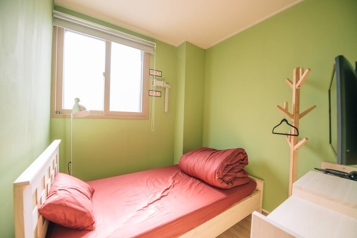 A single bed with a red mattress and pillows, a wooden nightstand, a window with blinds, a coat rack, and a television on a stand.