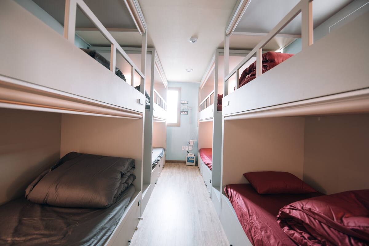 A narrow, well-lit room with bunk beds on either side, a small bedside table, and a window at the far end.