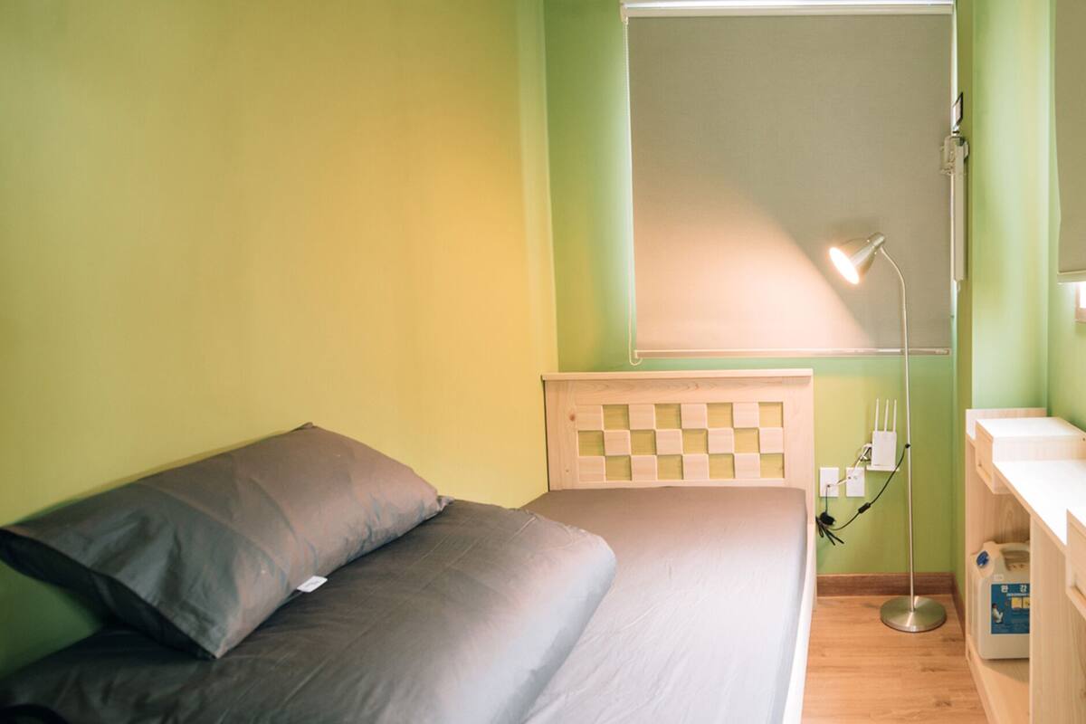 A small, neatly arranged bedroom with a bed, pillows, a bedside table, a lamp, and a window.