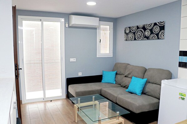 Una sala de estar moderna con un sofá gris, dos cojines azules, una mesa de centro de vidrio y un refrigerador blanco.