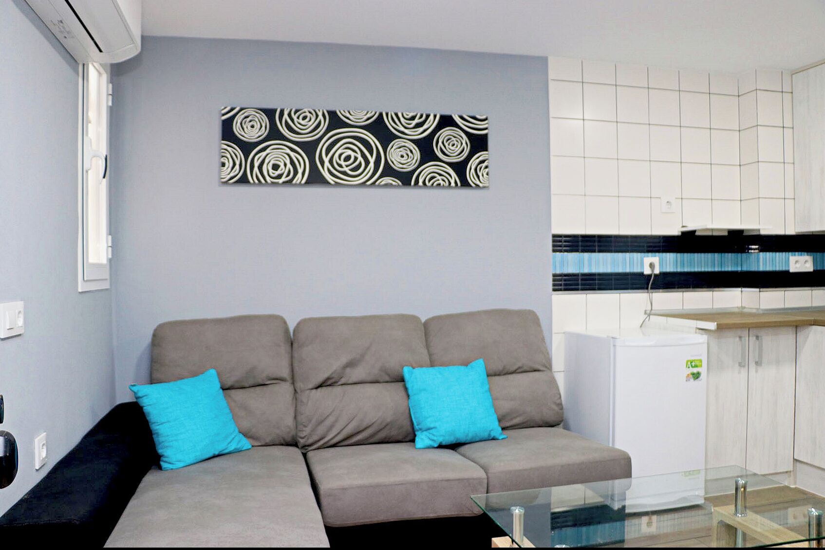 Een moderne woonkamer met een grijze bank, twee blauwe kussens, een glazen salontafel, en een keuken met witte kasten en een zwart aanrecht.