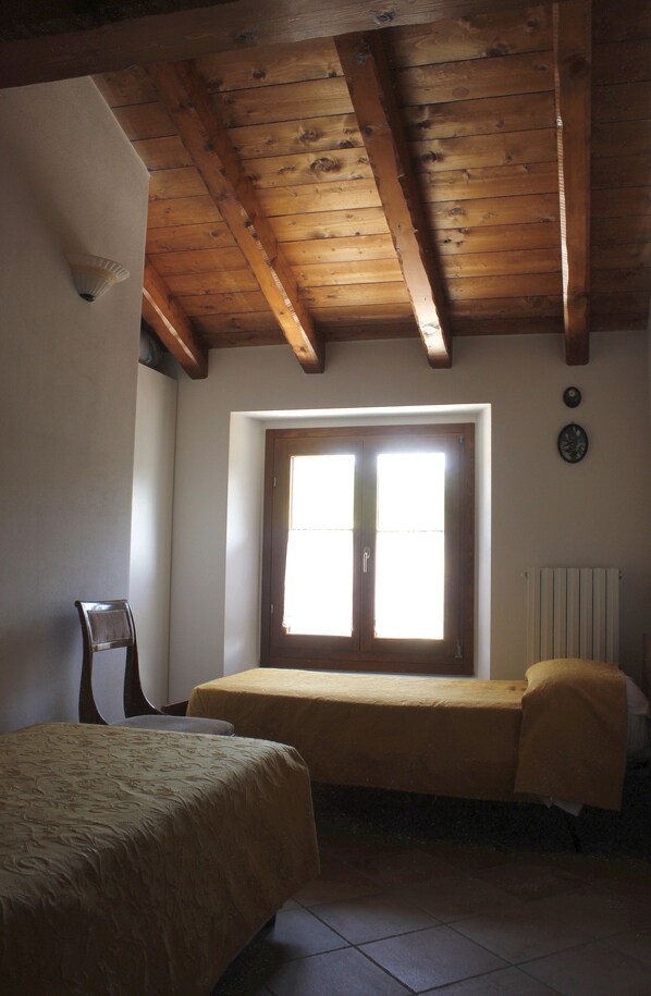 Una habitación con techo de madera, un ventanal con persianas, una cama con colcha amarilla, una silla y un radiador.