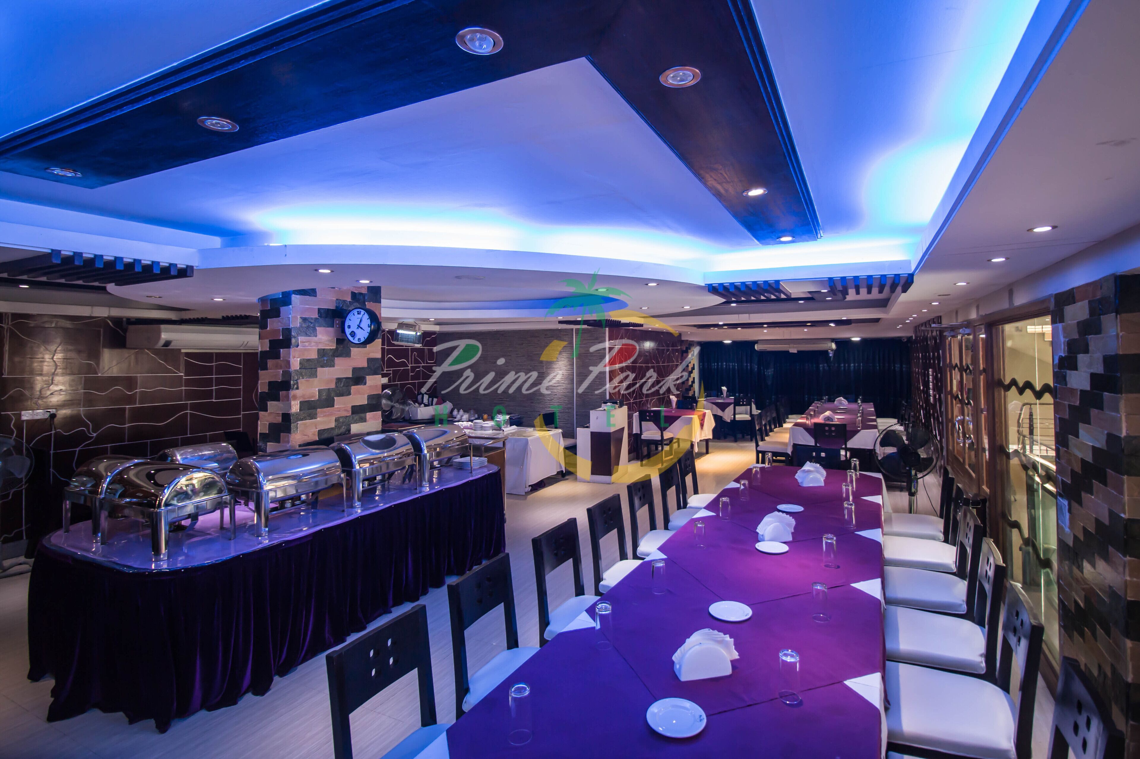 A restaurant with a long table set for dining, a buffet area, and a wall with a clock and palm tree decoration.