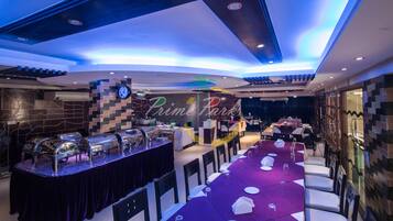 A restaurant with a long table set for dining, a buffet area, and a wall with a clock and palm tree decoration.