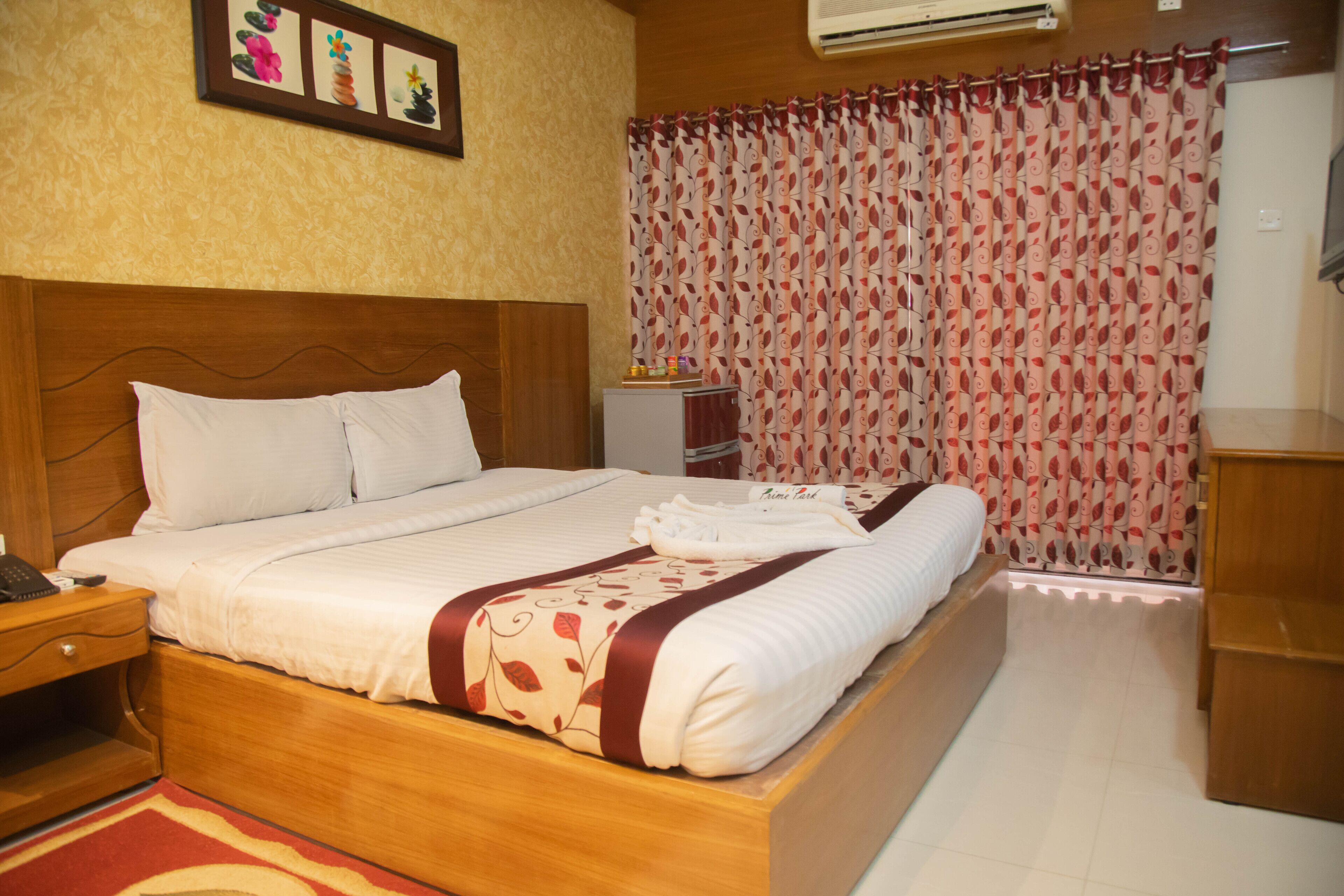 A neatly made bed with white linens and a decorative pillow, a wooden nightstand with a phone, a small cabinet, and a red and white patterned curtain.