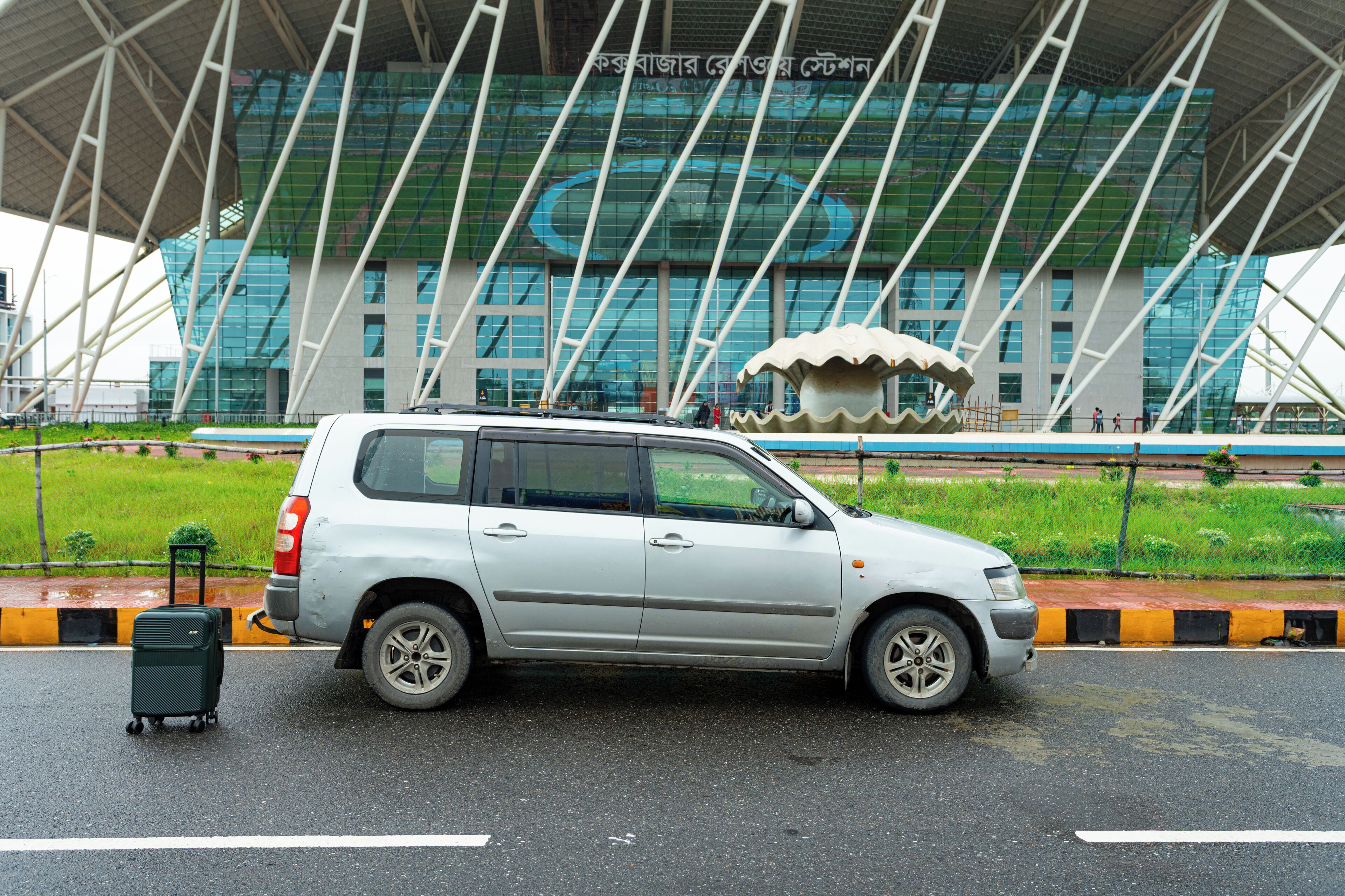 A silver SUV parked in front of a modern building with a distinctive roof structure and a large, decorative sculpture.