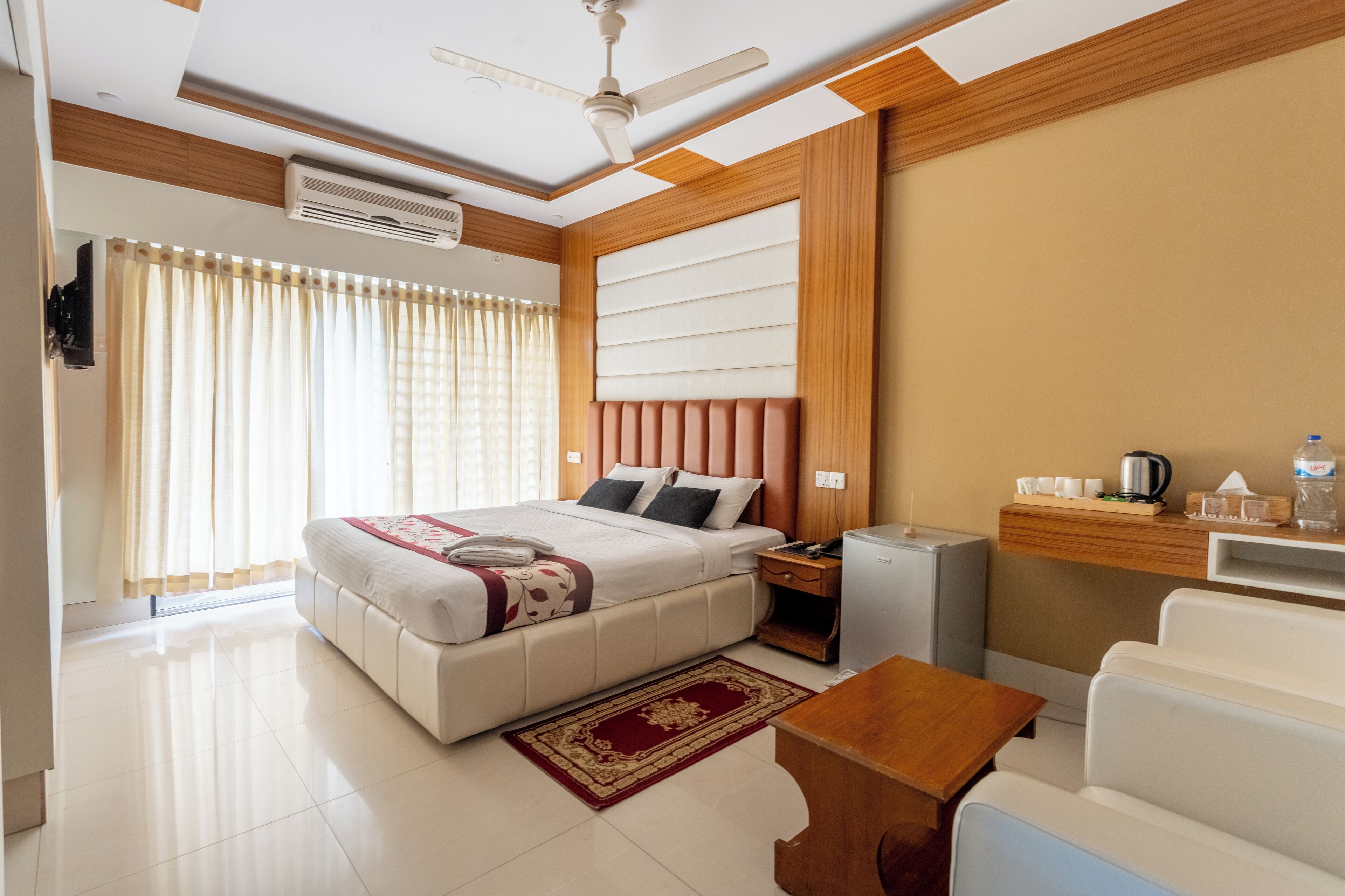 A modern hotel room with a large bed, a desk, a chair, a ceiling fan, and a small kitchenette.