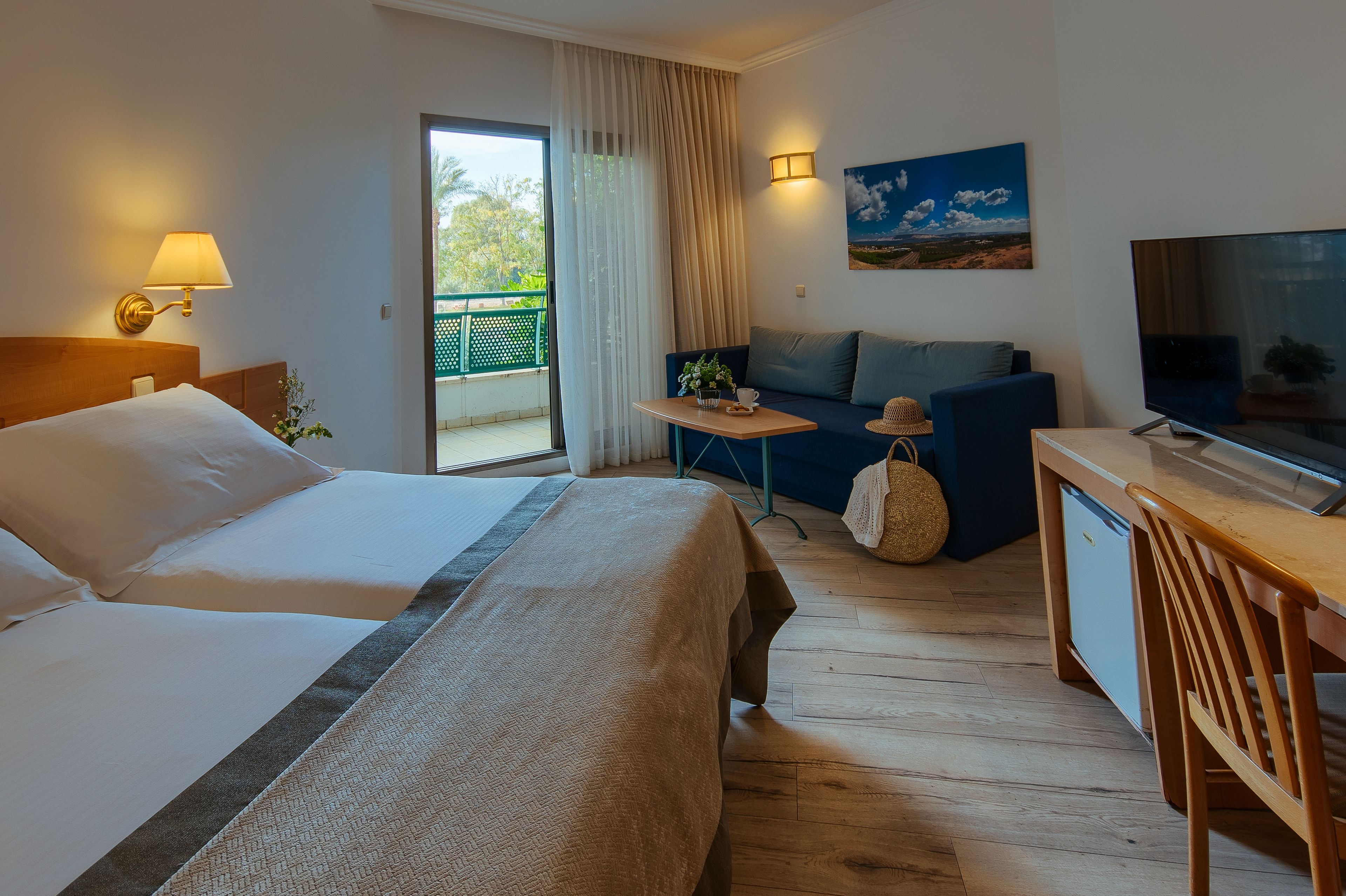 A hotel room with a bed, a sofa, a small table, a TV, and a balcony with a view of greenery.