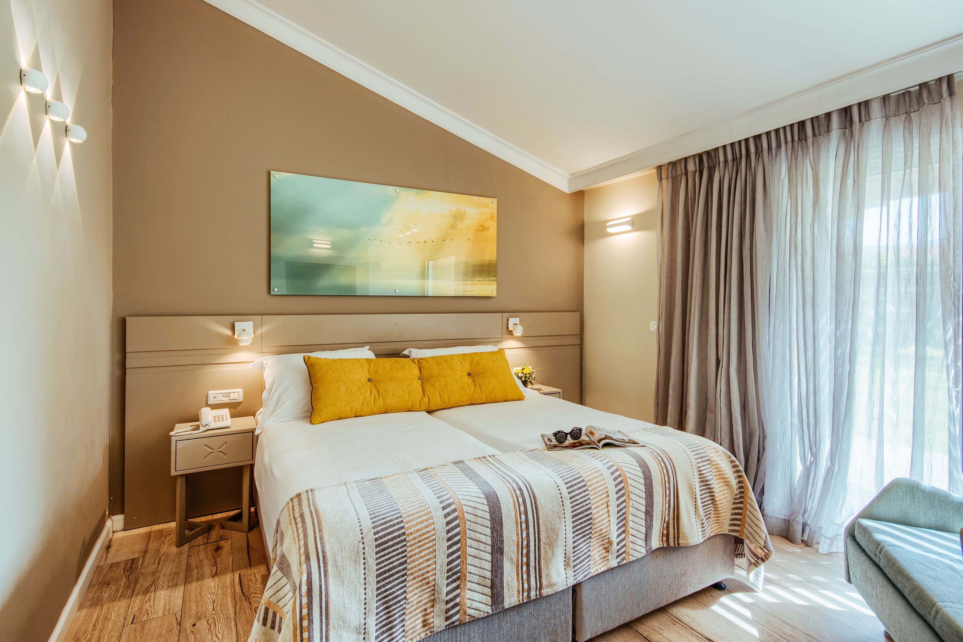 A hotel room with a large bed, a bedside table with a phone, a nightstand, a window with curtains, and a painting on the wall.