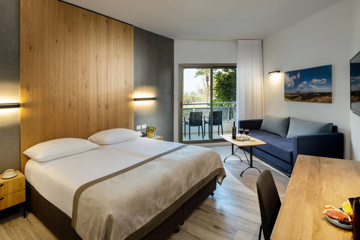 A modern hotel room with a large bed, a sofa, a dining table, and a balcony with outdoor furniture.
