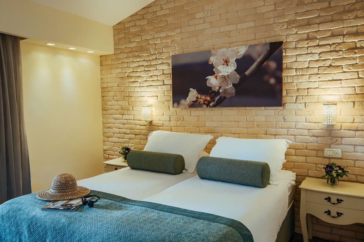 A hotel room with a brick wall, two beds with white linens, a blue textured bedspread, a hat, and a pair of glasses on the left side.