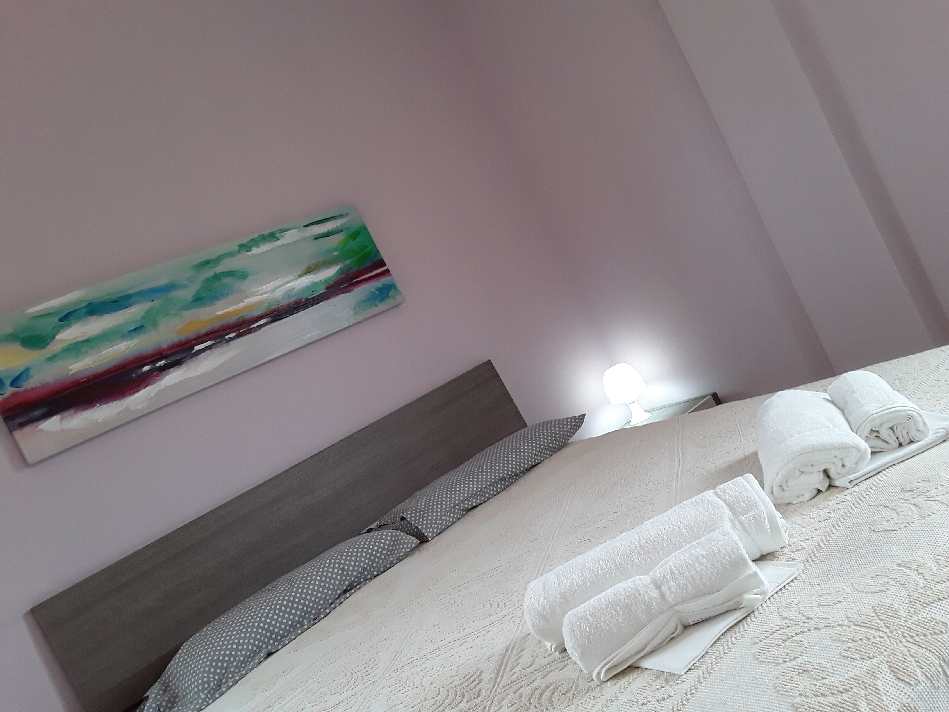 Uma cama bem arrumada com lençóis brancos e toalhas enroladas, um travesseiro cinza com bolinhas e um quadro de uma paisagem na parede.