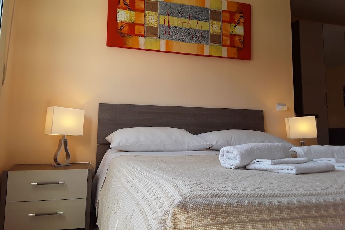 A hotel room with a bed, two bedside tables with lamps, a nightstand, and a colorful abstract painting on the wall.